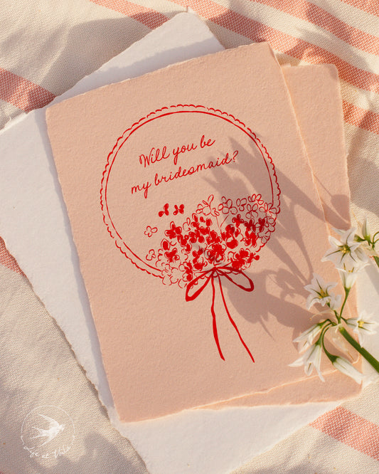 Card with 'Will you be my bridesmaid?' question and floral design on a soft fabric background.