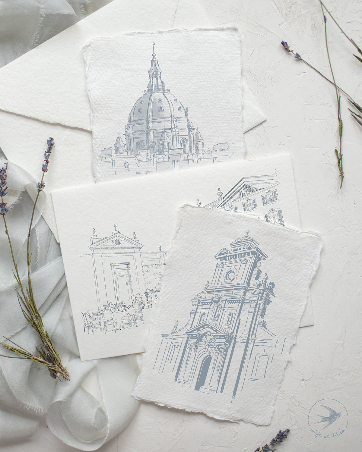 A collection of custom venue drawings displayed on a table, depicting various architectural styles, with lavender sprigs scattered around.